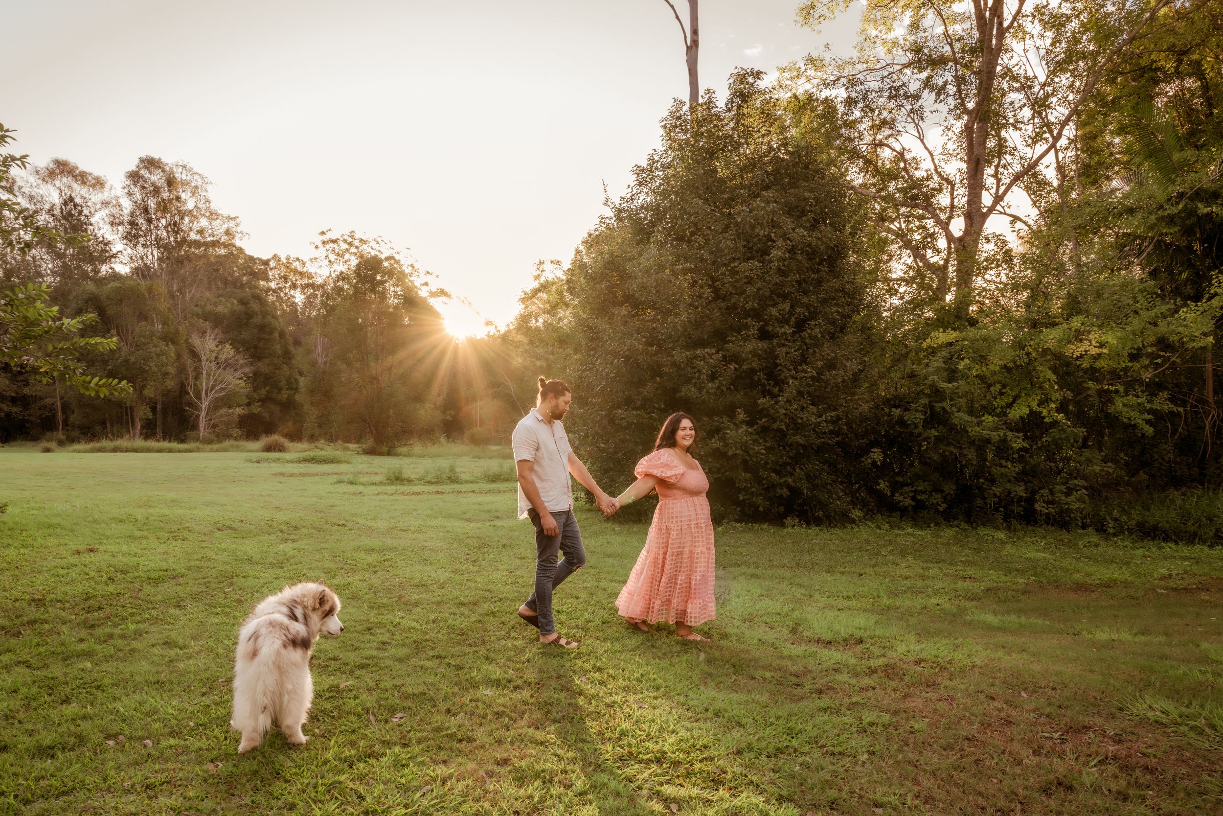 Couple holding hands in a sunlit green field at golden hour with a large white dog nearby.