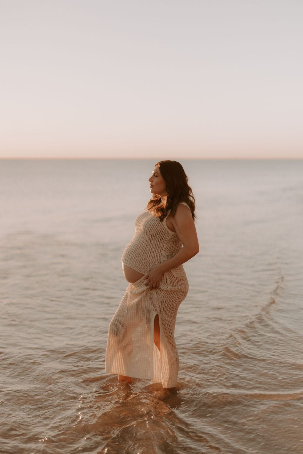A pregnant woman in a white dress stands in shallow ocean water during golden hour.