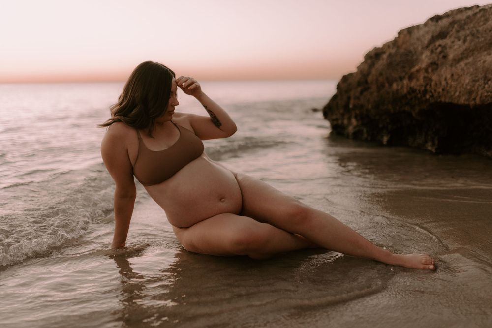 A woman poses on the beach at sunset in artistic maternity photos.