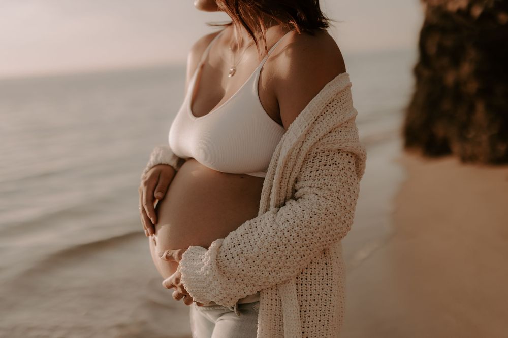 A maternity photoshoot at sunset shows an intimate moment on the beach with a white top and knit cardigan.