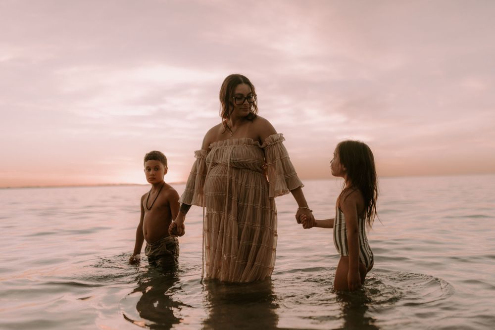 Family plays in ocean waves during sunset maternity photoshoot.