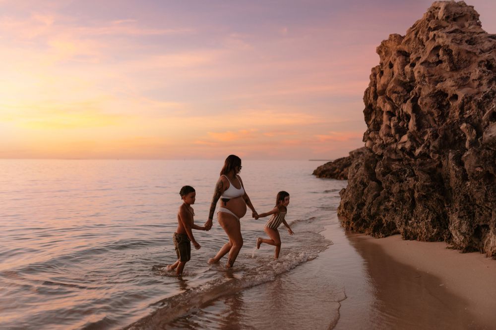 Silhouettes play in ocean waves against a pink sunset sky.