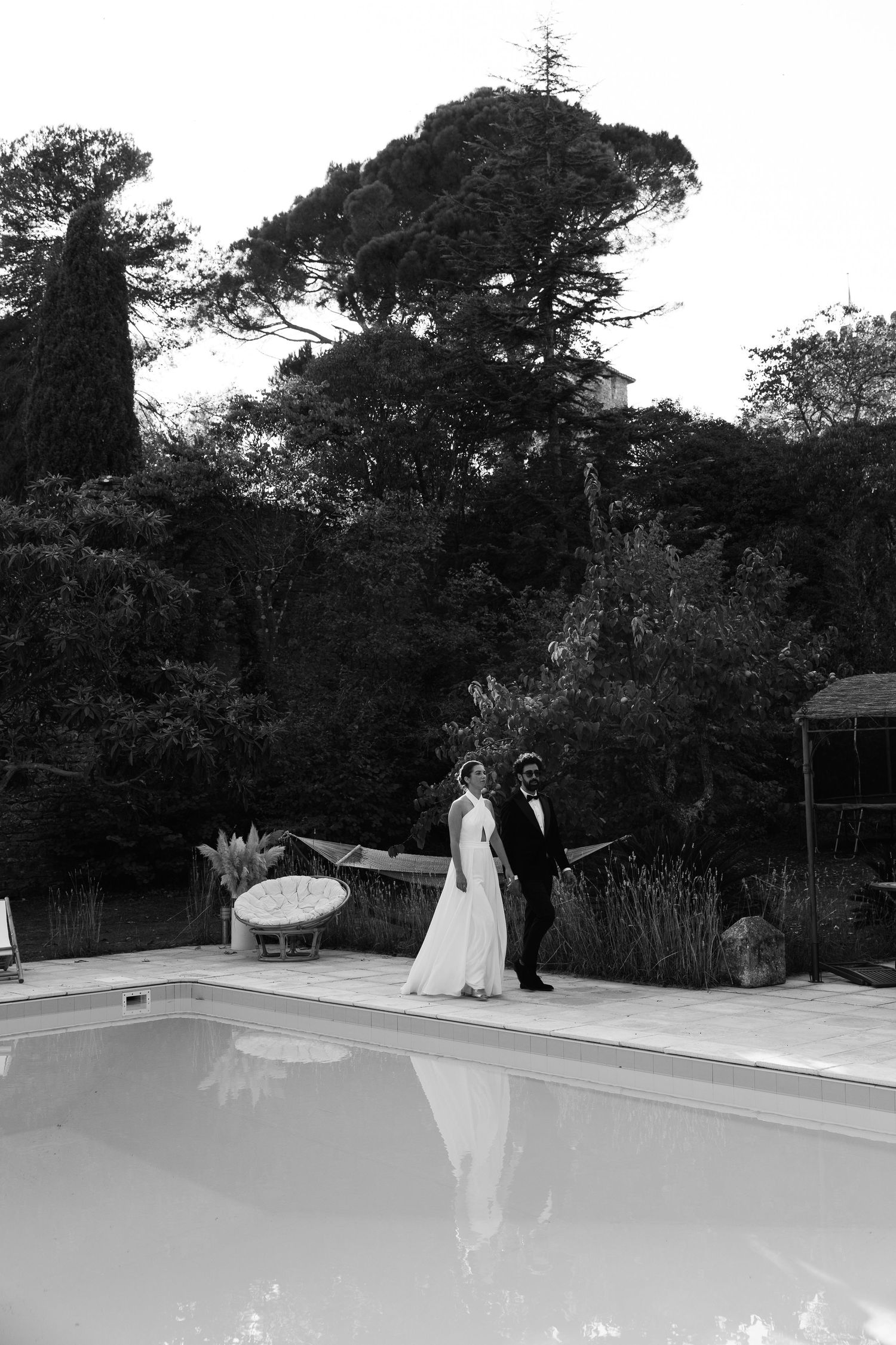 A couple stands together by a swimming pool surrounded by tall trees in black and white.