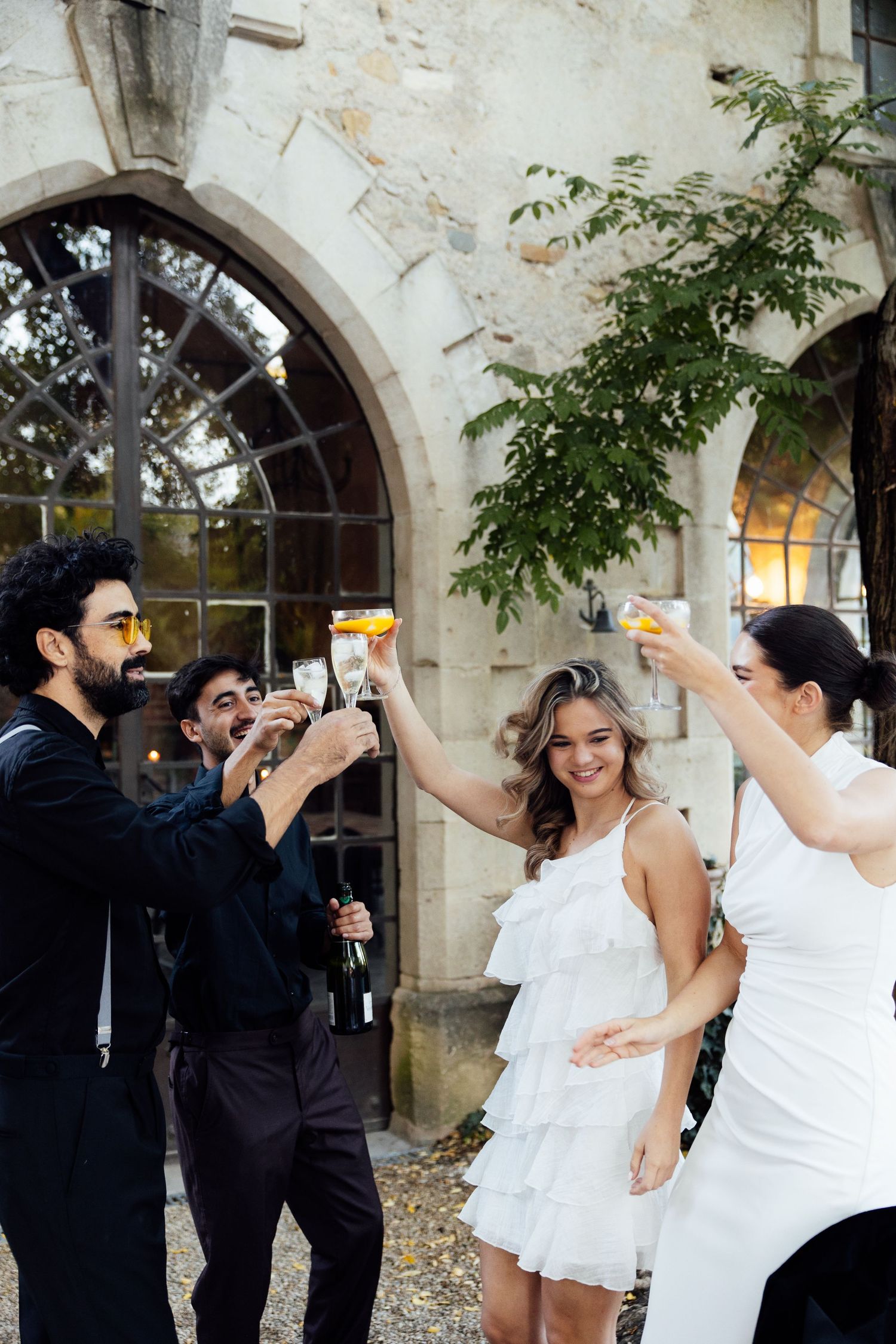 Wedding party in white dresses and dark suits dancing and celebrating at a rustic stone venue.