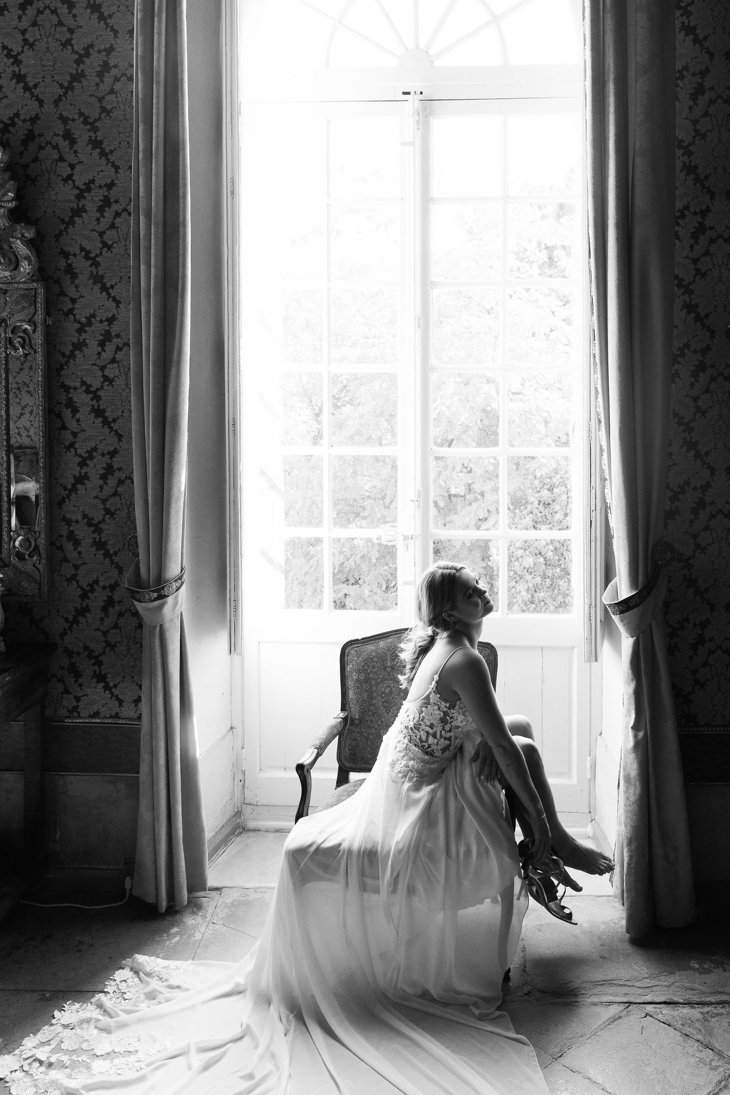 Black and white photo of a bride in flowing gown sitting by a bright window in an elegant room with ornate wallpaper.