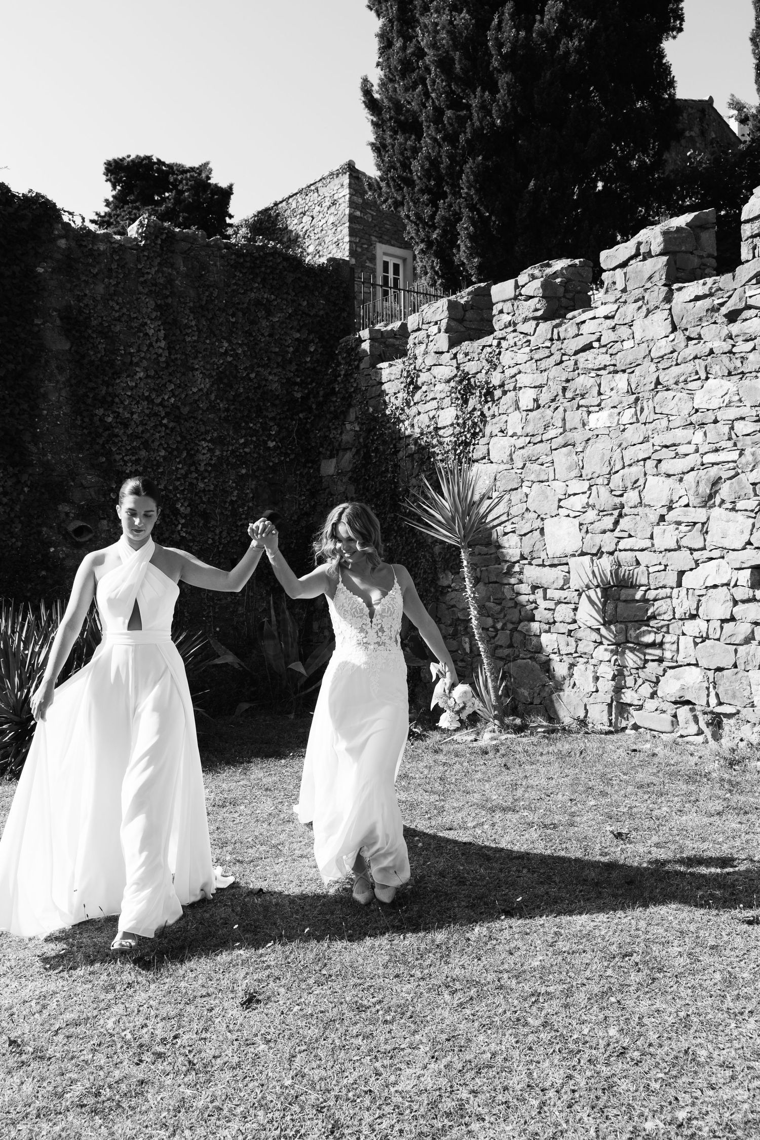 Two people in flowing white dresses hold hands while walking on grass near a rustic stone wall.