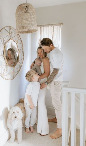 Family portrait in white room with geometric mirror and woven pendant light, dog sitting nearby.