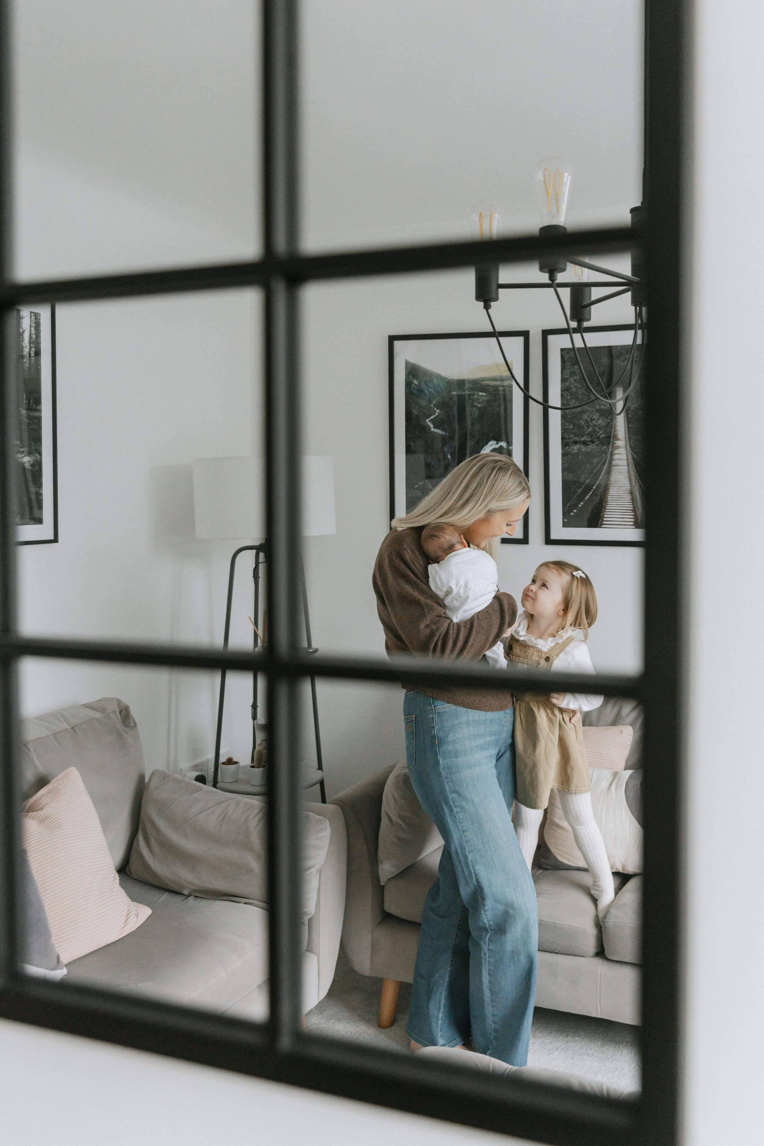 Three window panes showing a person in denim overalls arranging decor on a white wall above a gray couch.