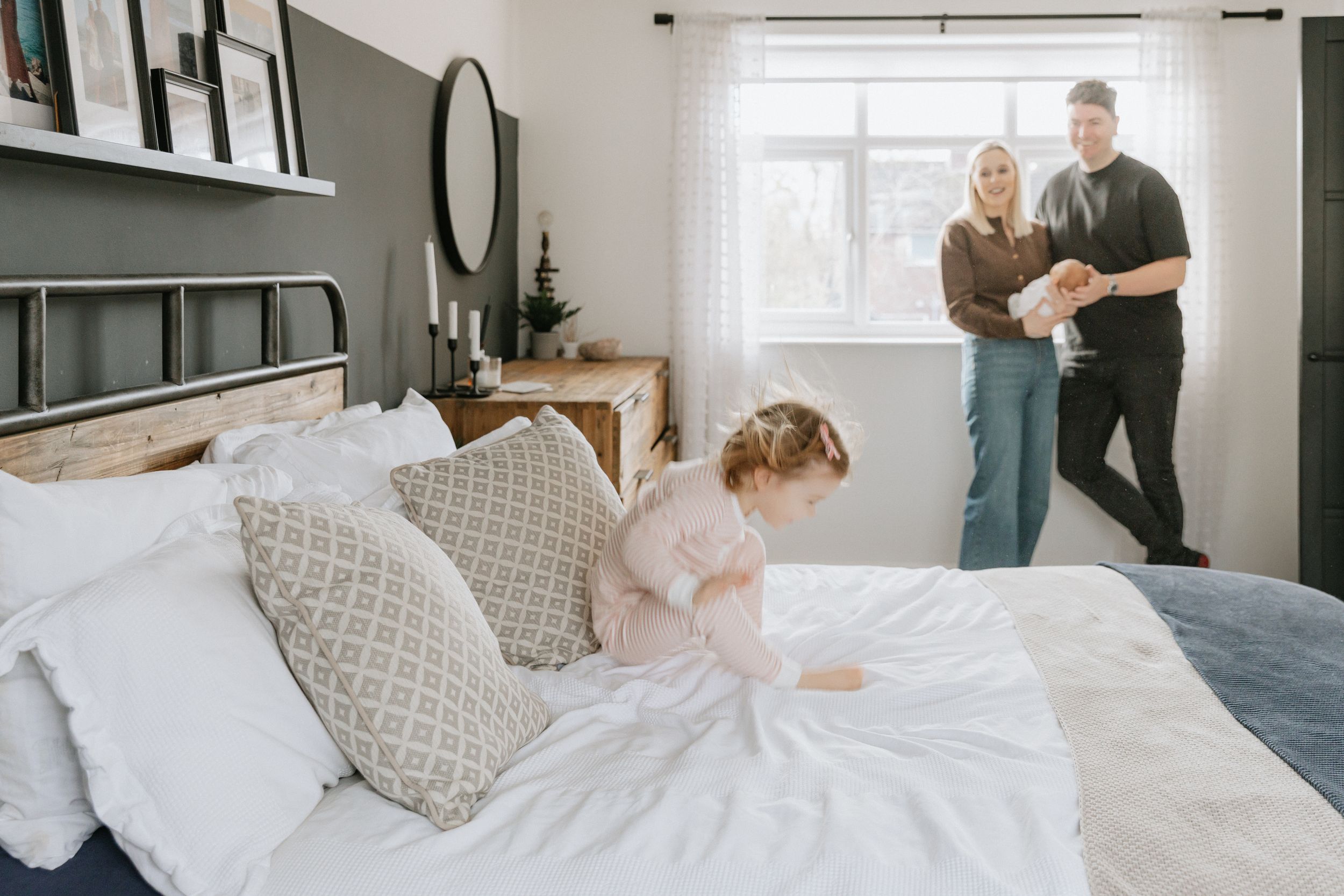 A bright modern bedroom features white bedding, geometric pillows, and natural light streaming through windows.