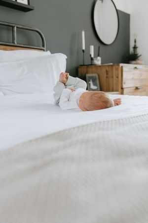A small baby in light blue sleepwear rests peacefully on white bedding in a minimalist bedroom with sage green walls.