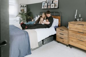 A modern bedroom with dark gray walls features a wooden dresser, cozy bedding, and natural light from windows.