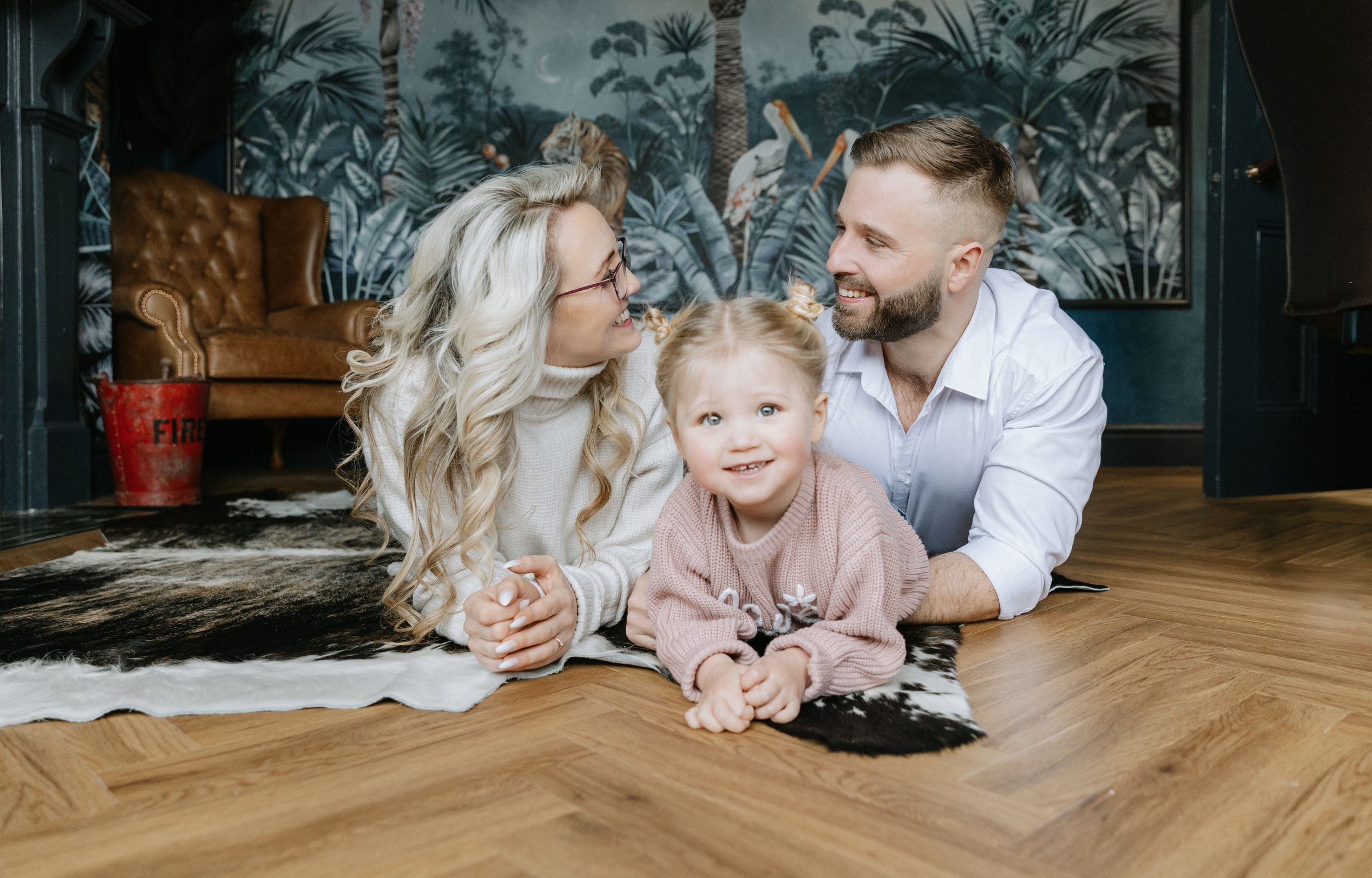 A family lying on hardwood floor together with tropical wallpaper and leather chairs in background.