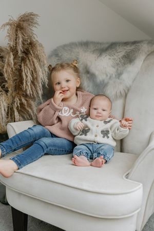 Two siblings sitting on white armchair with pampas grass decoration.
