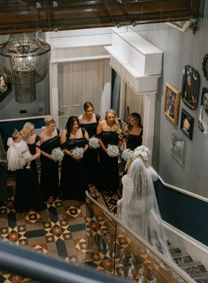 Bridesmaids in black dresses gather on a staircase with ornate flooring and a crystal chandelier during a wedding celebration.