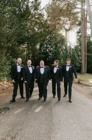 A group of groomsmen in black tuxedos walk together on a gravel path surrounded by trees.