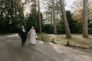 A couple walks together through a scenic wooded park path during a formal occasion.
