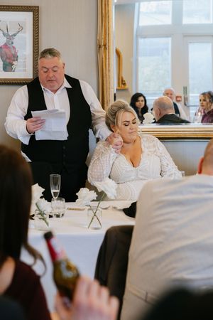 Couple at a formal reception with table settings and champagne glasses while someone reads from a paper.