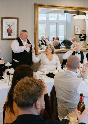 Guests in formal attire gather at tables during an elegant dinner event in a bright dining room.