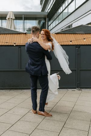 A couple in wedding attire dances and twirls together on a modern rooftop terrace with glass walls in the background.