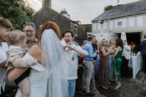Wedding guests gather outside a rustic stone building during a sunny outdoor celebration.