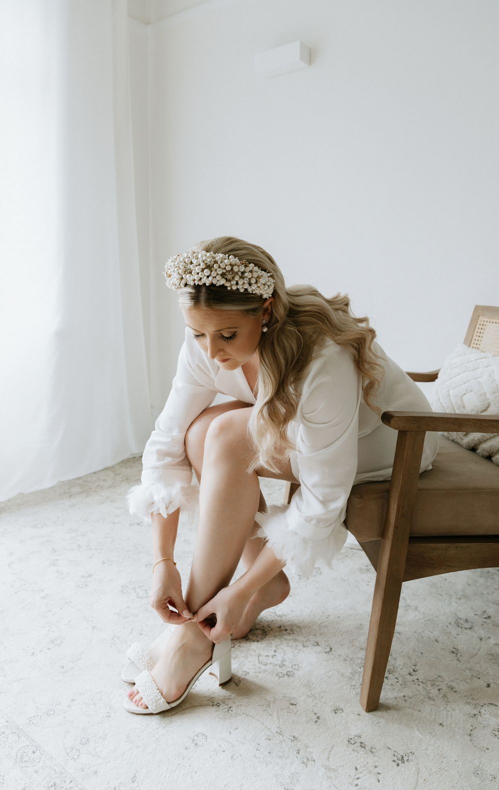 Close-up of a bride in white dress fastening elegant wedding shoes while sitting on wooden chair.