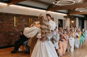 Wedding couple performs their first dance while guests in pastel dresses watch from a line against exposed brick walls.