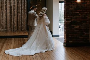 A bride in a flowing white wedding gown twirls gracefully on a hardwood floor in an industrial brick venue.