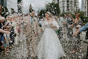 Wedding guests toss confetti over a couple in formal attire outside a grand building.