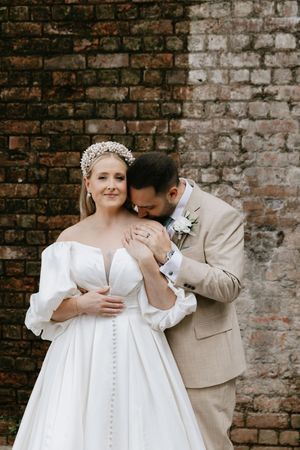 Romantic wedding couple poses against rustic brick wall in elegant white dress and beige suit.