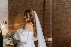 A bride in a white dress stands near exposed brick walls with natural window lighting creating a soft glow.