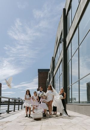 A group in white dresses stands on a modern rooftop terrace with glass windows and blue sky in the background.