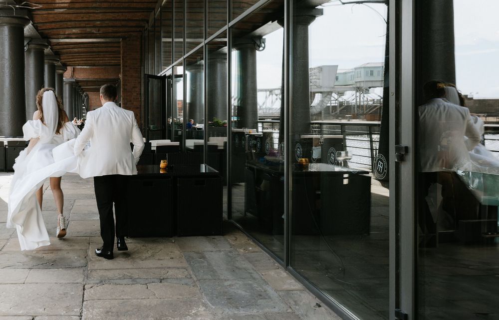 A wedding couple stands at a modern restaurant bar with floor-to-ceiling windows overlooking a city view.