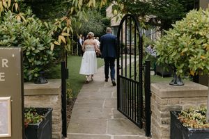 A couple walks down a garden path between black iron gates surrounded by lush greenery and flowers.