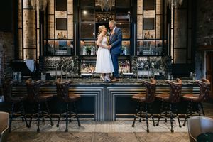 A romantic wedding couple shares an intimate moment in front of an industrial brick building with string lights.