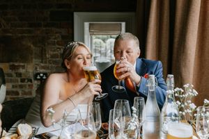 Wedding guests sit at long tables during a reception dinner with wine glasses and place settings in a rustic venue.