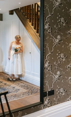 Person in white dress examines ornate patterned carpet near floral wallpaper and wooden staircase in elegant interior.