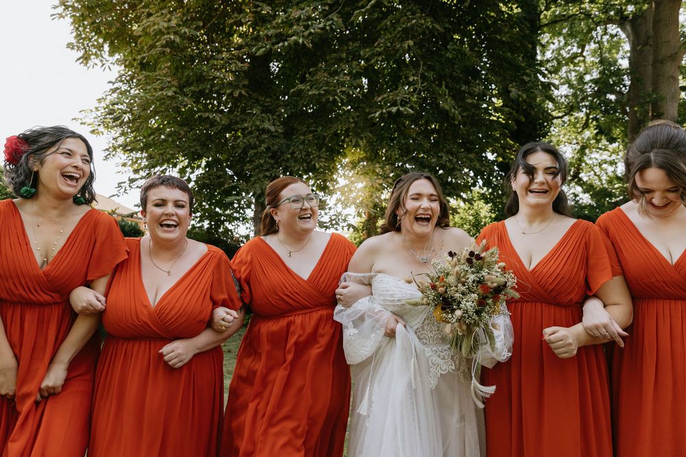 A group of bridesmaids in matching red dresses walk arm in arm while laughing together outdoors.
