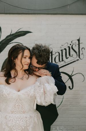 A romantic wedding moment captured against a white brick wall with vintage lettering, featuring a lace dress and navy suit.