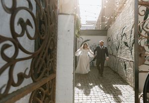 A couple walks down a narrow cobblestone alley with ornate wrought iron railings and stone walls.