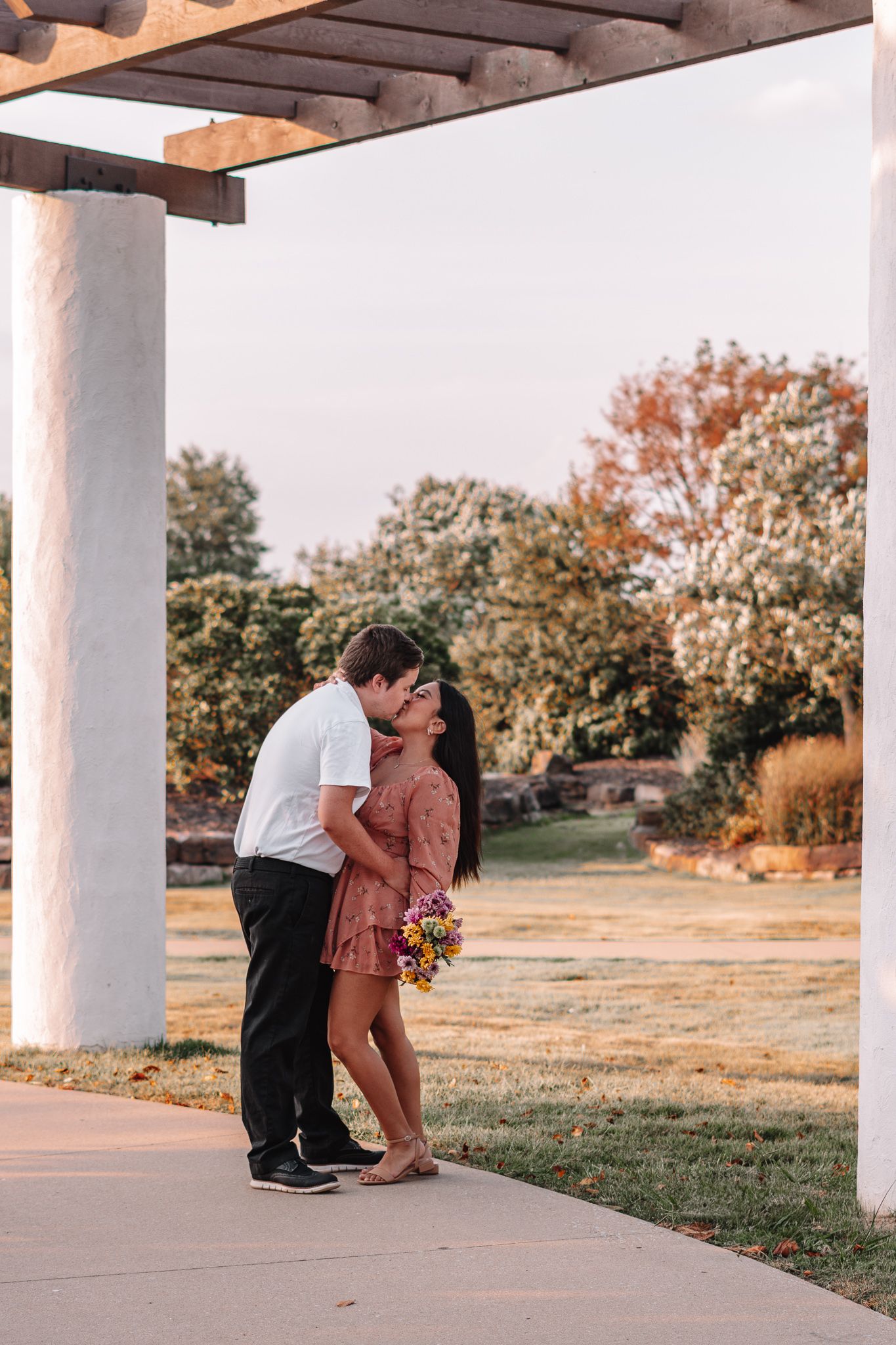 Couple embracing under white pillars during sunset with a scenic outdoor backdrop.