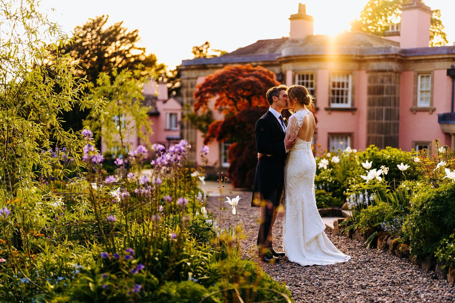 Bride and groom share a romantic kiss in a lush garden at golden hour, with a pink manor house in the background.