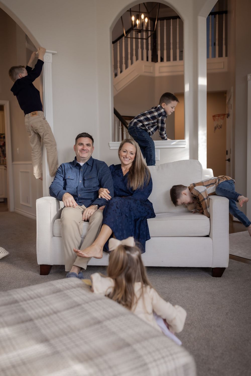A happy couple sits on a white sofa smiling while their energetic children play around them in a bright, elegant home.