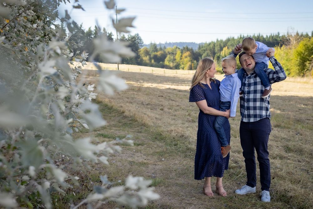 Family of four posing outdoors in a sunny field, parents embracing children with trees and hills in the background.