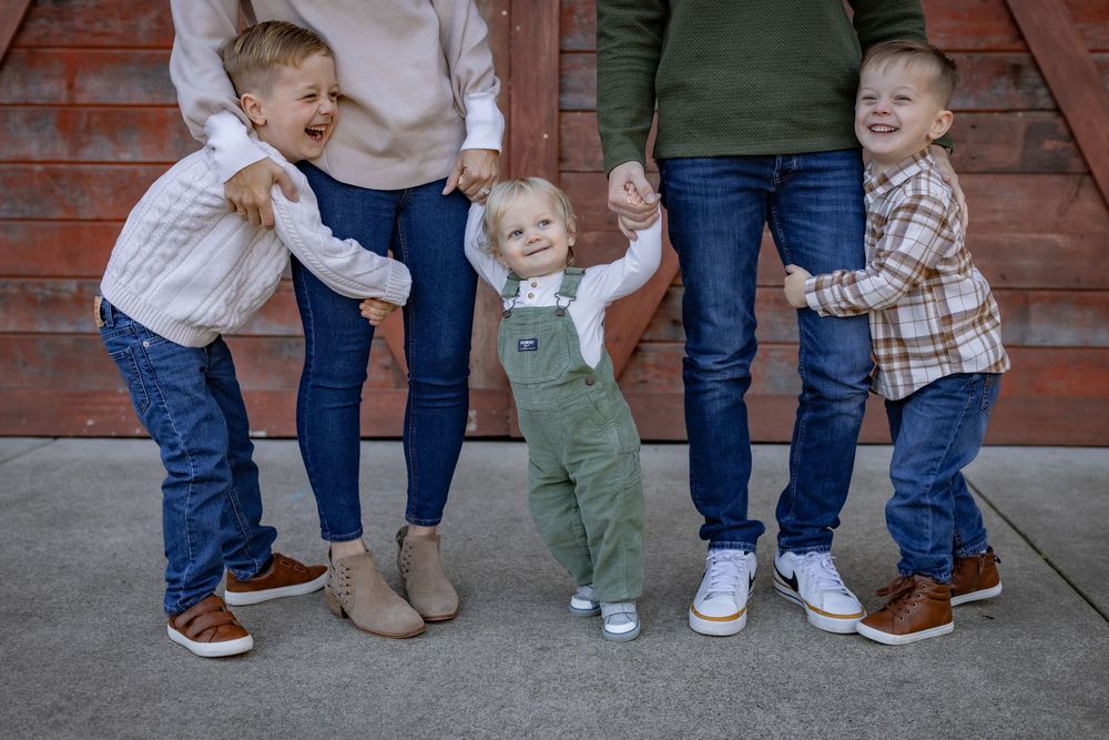 Family with two toddlers and a baby holding hands outdoors, wearing casual fall outfits in front of a red barn.