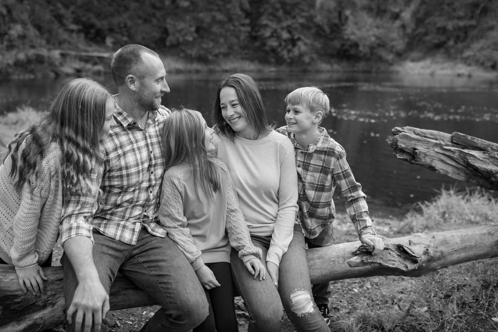 Black and white family photo of parents and two children sitting on a log near a river in a forest setting.