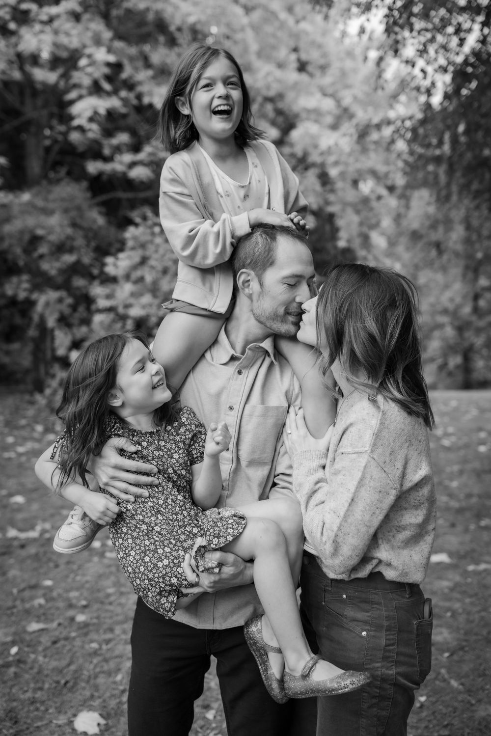 Black and white family portrait of parents with two young daughters laughing outdoors in a park setting.