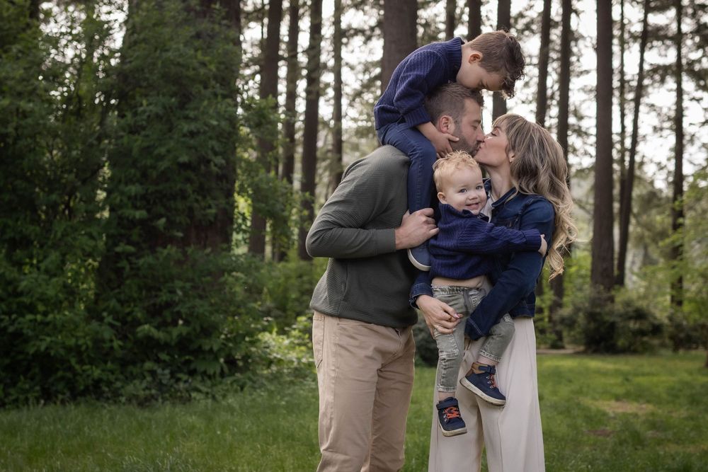 Family of four sharing a tender moment outdoors, parents kissing while holding two young boys in a lush green forest setting.