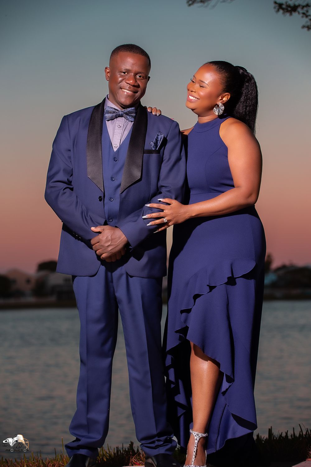 A couple in matching navy formal wear poses at sunset by the water.