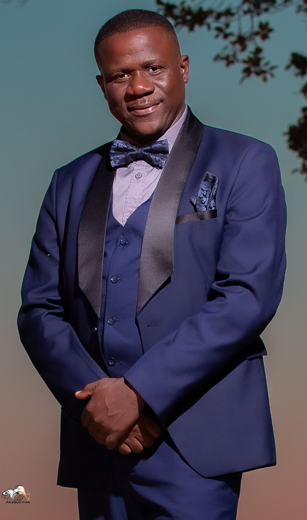 A person in a navy blue three-piece tuxedo with bow tie poses against a sunset backdrop.