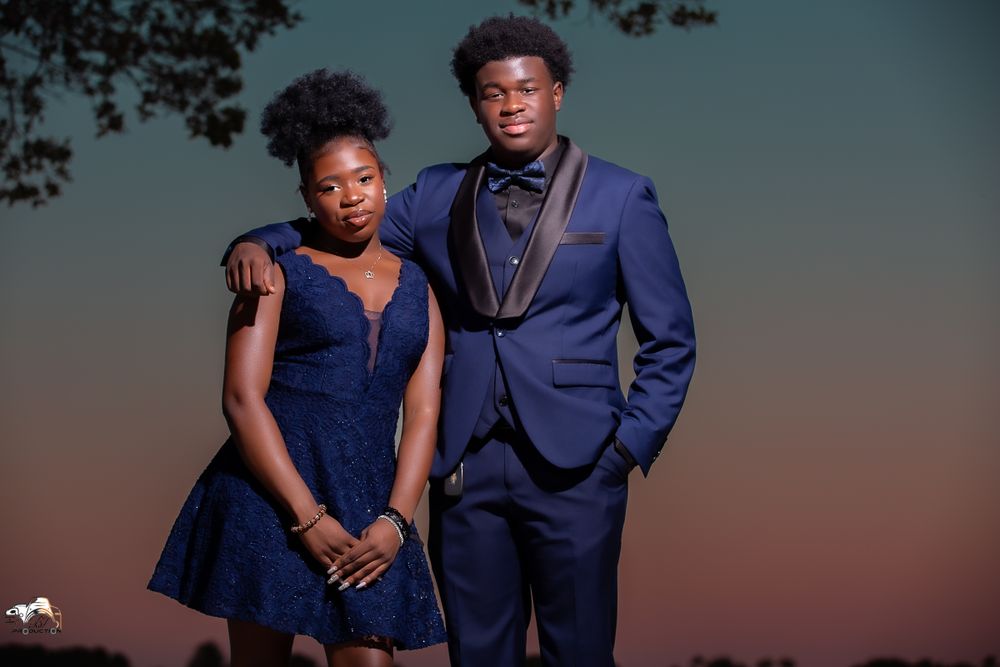 A couple in coordinating navy formal wear poses against a dusky sunset sky.